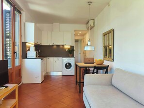 The living room features a comfortable-looking light-colored sofa positioned in front of a wooden table. The kitchen is visible in the background, equipped with a washing machine and modern white cabinets. The apartment has terracotta tile floors througho