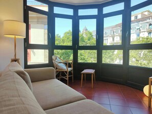 The living room is bathed in natural light streaming through a large bay window offering stunning views of the city. A comfortable couch sits in front of the window, inviting you to relax and soak in the sights. A wicker chair and a stool provide addition