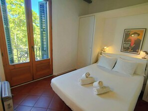 The bedroom is brightly lit and features a comfortable queen-sized bed with crisp white linens and fluffy towels neatly folded on the bedspread. The room boasts a large window with wooden shutters offering views of the leafy trees outside. A vintage poste
