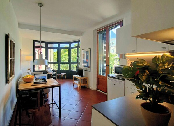 The open-plan living space features a large bay window offering natural light and views of the bustling La Rambla. A dining table sits near the window, ideal for enjoying meals while watching the street scene below. The apartment is tastefully decorated w