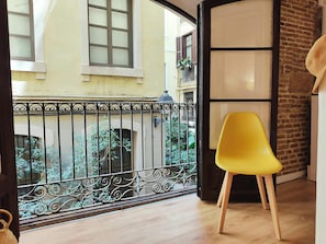 The loft offers a glimpse into the heart of Barcelona overlooking a quiet, private passage. Natural light illuminates the stylish wooden floor and highlighting the bright yellow chair situated near the window. The wrought-iron railing provides an elegant 