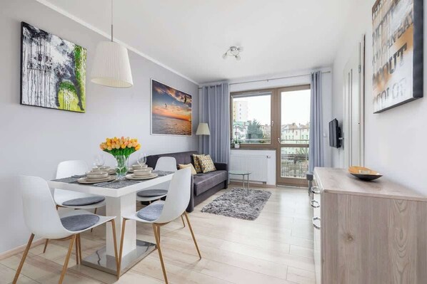 A dining area in a modern apartment with a round white table, four white chairs, and flowers as a centerpiece. Large windows provide natural light.