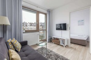 A bright living room with a comfortable gray couch, decorative cushions, and a large window overlooking the city. On the wall is TV.