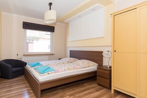 A bright bedroom with a double wooden bed and a comfortable mattress next to which stands a yellow wardrobe on one side and a pouffe on the other. 