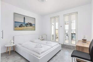 A bedroom with a large bed covered in crisp white linens, a sleek modern dresser, and access to a balcony or terrace with plenty of natural light streaming in.