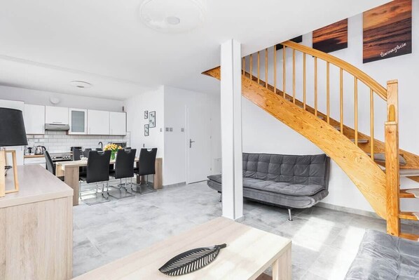 The living room is bright and spacious, featuring modern furniture, including a large sofa and a wooden staircase that adds a touch of warmth to the space. Large windows provide ample natural light.