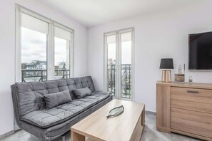 The living room offers a cozy space with a gray sofa, a large window, and a flat-screen TV mounted on a light-colored wall, creating a relaxing area for entertainment.