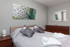 A cozy bedroom featuring a large bed with multiple pillows and decorative art above the bed. There is also a wooden dresser with a mirror.