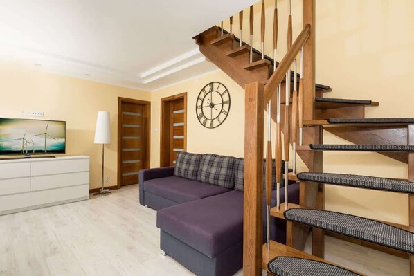 Bright, spacious living room with a chest of drawers on which stands the TV. To the right is a corner sofa bed above which is a village clock. In the right corner there are stairs leading upstairs.
