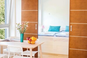 A cozy bedroom with a neatly made bed, featuring wooden wall panels and bright natural light from a window. A small bedside table holds a potted plant, adding a touch of nature.