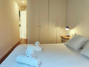 This is a photo of the queen-size bed in one of the bedrooms of the apartment. The bed is made with crisp white linens and fluffy white pillows. There are also four white towels neatly rolled up on the bed, which are included for guest use. The bedroom fe