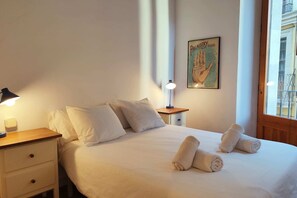 The bedroom features a comfortable queen-size bed with crisp white linens and fluffy pillows. The bedroom is bathed in natural light, streaming through a nearby window with wooden shutters.  Two rolled-up white towels are placed neatly on the bed, a welco
