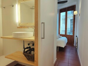 The bathroom features a modern, white sink and sleek chrome fixtures, with a wooden shelf underneath for storage. A wicker basket sits on the lower shelf, perfect for storing toiletries.  The bathroom is connected to a bedroom with a double bed, visible t