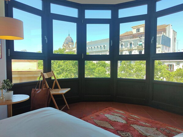 The apartment features a large bay window offering panoramic views of the city, including the iconic rooftops of Barcelona. The window is framed by black wooden molding and allows natural light to flood the room, creating a bright and airy atmosphere.  Th
