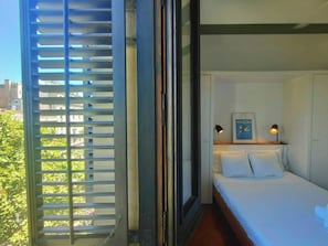 This photo shows the bedroom of the apartment. The bedroom features a large window with wooden shutters and a view of the street below.  The bed is made with crisp white linens and there are two bedside lamps with black shades.  The apartment is located o