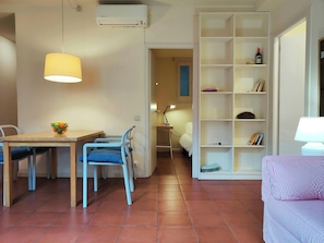 Spacious living room. A large white bookcase with various books.  The bookcase is adjacent to a doorway, allowing a glimpse of a second room with a bed, a desk, and a lamp. The room has a warm, inviting feel with terracotta tile floors and bright, natural