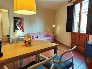 The spacious living area features a large wooden dining table with four white chairs. A pink striped couch sits to the right of the table. A large window with wooden shutters and a view of the street is on the right side of the room. A bright yellow lamp 