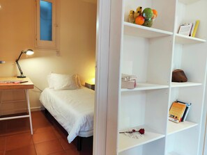 The second bedroom features a twin bed with crisp white linens and a bedside lamp. The room is bright and airy with a small window, providing natural light. A desk with a lamp is situated next to the window, offering a space for working or reading. The ro