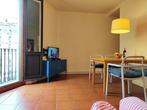 The living room features a dining table with four chairs, a television mounted on a blue cabinet and a large window overlooking a traditional Barcelona street. The tile flooring adds a touch of classic charm to the space.