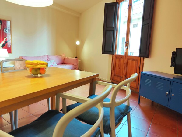 Inviting living space, surrounded by comfortable white chairs with blue cushions. The room also features a striped sofa, a flat-screen TV mounted on a stylish blue cabinet, and a view of the street through a set of French doors with wooden shutters. This 