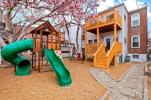 Fenced in playground for kids in the backyard with two slides, rock wall & more.