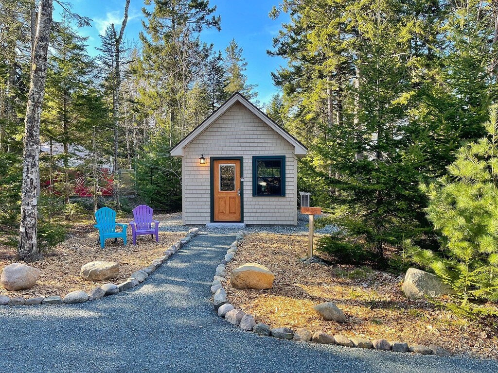 A tiny house in Acadia National Park sits at the end of a long walkway with two colorful adirondack chairs beside the path, and trees completely surrounding.