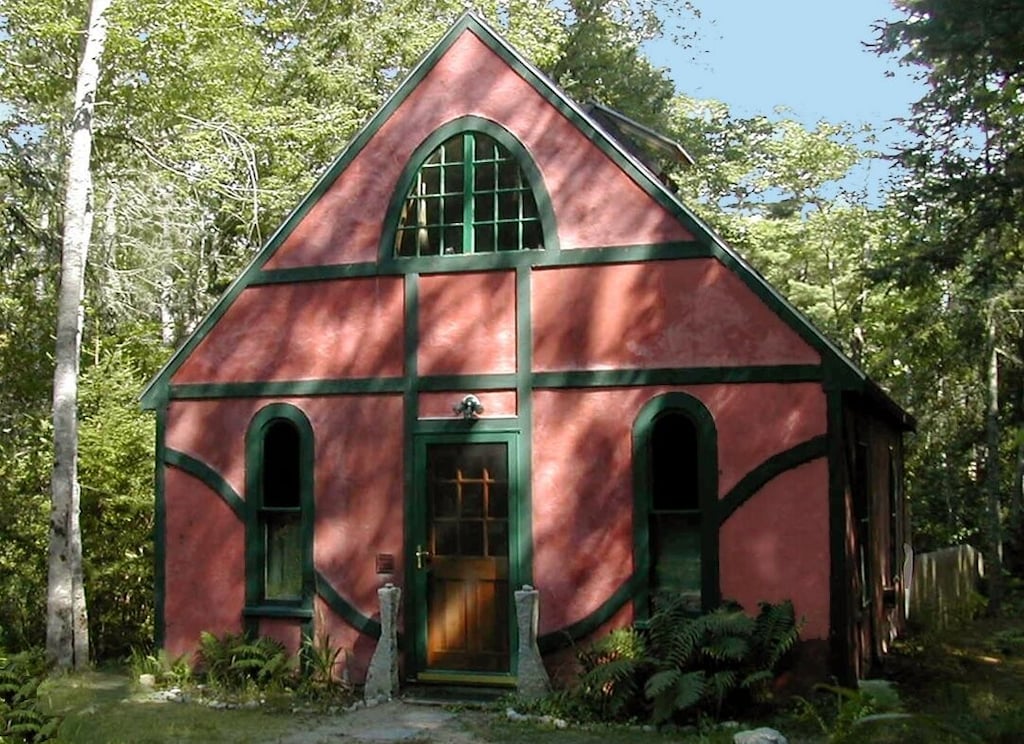 Cute cottage vacation rental with red walls and green trim is seen with arched windows and surrounded by green grass and trees.