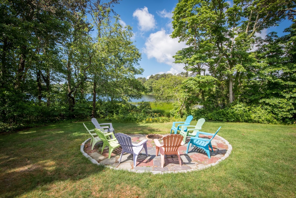 A fire pit with ample seating is seen surrounded by trees on a grassy spot with water in the background