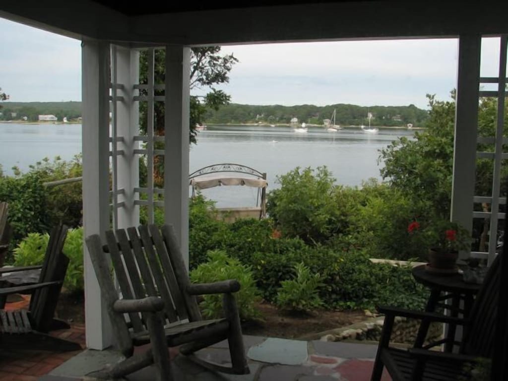 A patio with two rocking chairs sits surrounded by green foliage with a body of water beyond