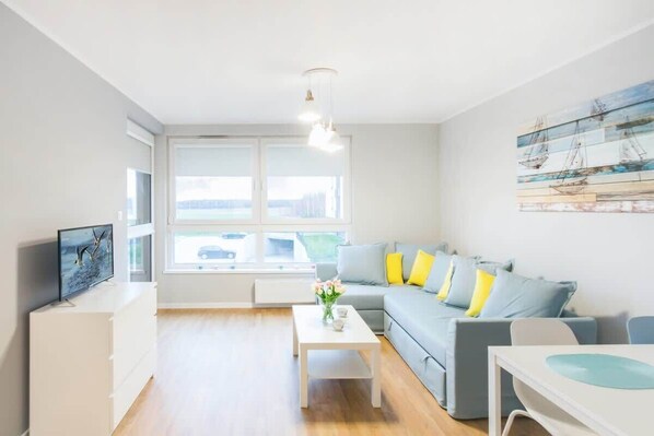 A bright and modern living area featuring a large sofa with colorful cushions, a coffee table, and a wall-mounted TV. Large windows allow plenty of natural light, creating a fresh and welcoming atmosphere.
