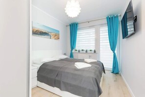 A bright bedroom with a large bed, neutral bedding, and a pop of color from turquoise accents. The room has ample natural light and a clean, minimalist design.