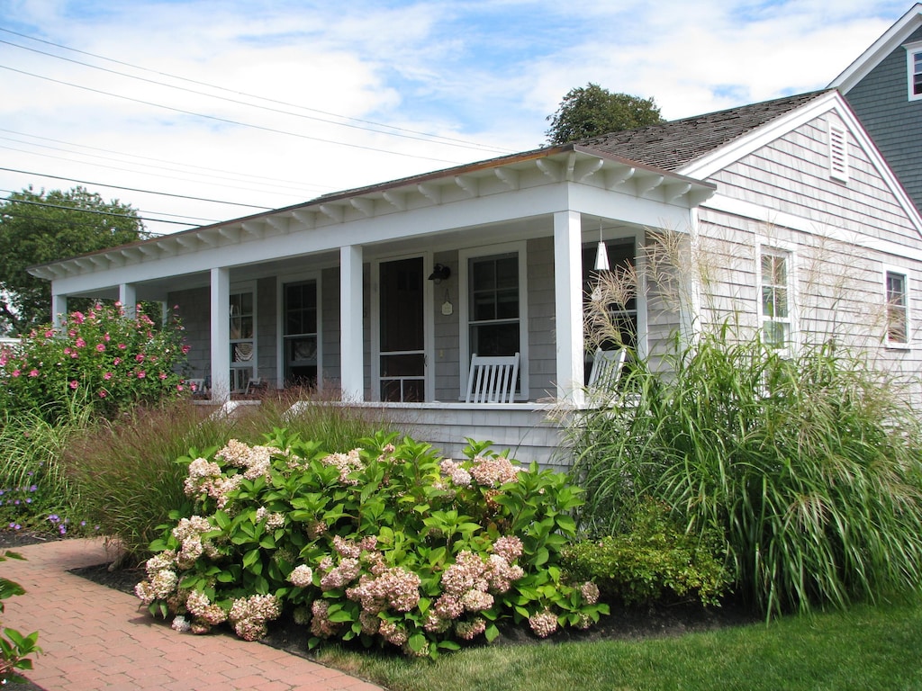 A grey Rhode Island summer cottage is seen with blooming flowers and grasses surrounding, a covered front porch, and blue skies above.
