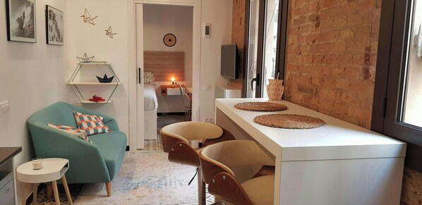 This is a view of the living room, featuring a light blue sofa with patterned cushions and a white side table with a candle. The room is tastefully decorated with minimalist furniture and an exposed brick wall.  A doorway leads into the bedroom, which fea