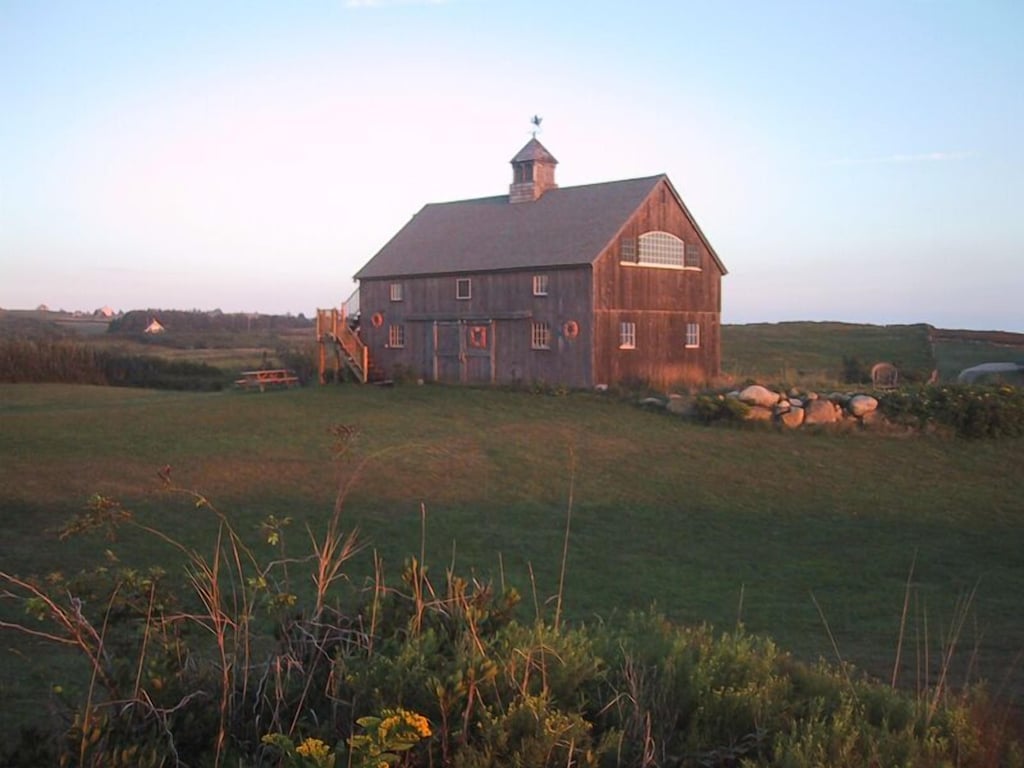 An idyllic barn vacation rental on Block island is seen with grass all around it and a soft sunset beyond