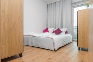A minimalist bedroom with a large bed dressed in white bedding and accented with purple pillows, creating a vibrant yet calming atmosphere.
