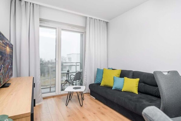 A bright living room with a light wood floor, cozy gray sofa, and large windows, creating an open and airy ambiance.