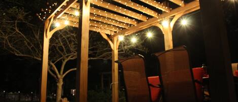 Pergola with fireplace, grill, patio furniture, lighted at night.