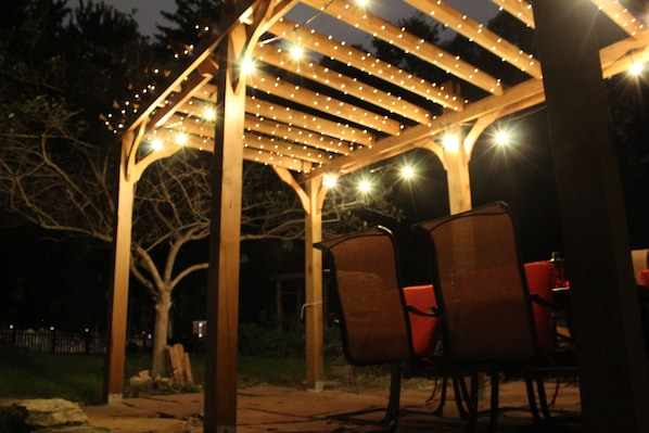 Pergola with fireplace, grill, patio furniture, lighted at night.