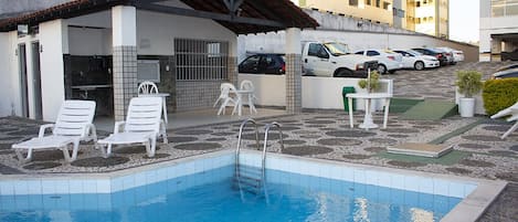 Área da piscina com: churrasqueira, banheiros, purificador de água, cadeiras para tomar sol, parque infantil, quadra de futebol infantil e chuveiro.