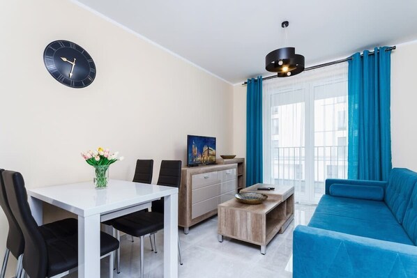  A living room with a blue sofa, dining table with chairs, and bright curtains. The space is lit with natural light from large windows.
