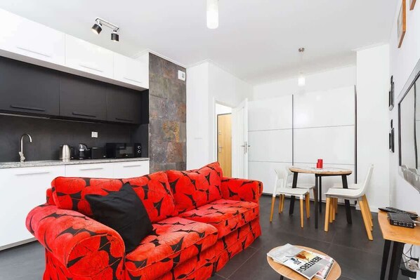 he first photo shows a living room combined with a kitchenette, featuring a patterned red sofa in the living area and white cabinets in the kitchen area.