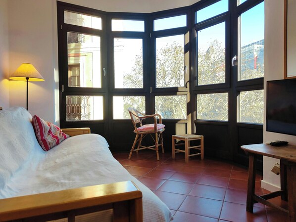 The living room features large windows overlooking the bustling streets of Barcelona. The view from the couch includes a glimpse of the trees and buildings beyond the window. The room is furnished with a comfortable sofa and a wicker chair, suggesting a r
