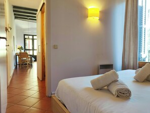 The bedroom features a comfortable queen-sized bed with crisp white linens, inviting you to relax after a day exploring Barcelona. The room is bathed in natural light streaming in from a large window with a light-colored curtain, creating a bright and air