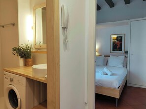 The bathroom features a modern white sink and a large mirror. It also includes a washing machine for added convenience. A glimpse of the bedroom is visible through the doorway, showcasing a neatly made bed. This image provides a peek into the functionalit