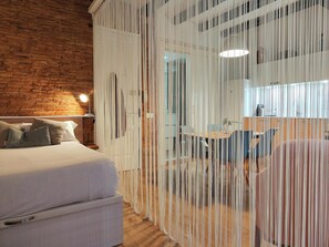 The inviting bedroom features a comfortable queen-size bed with storage space underneath, perfectly positioned against a rustic brick wall. A unique string curtain separates the bedroom from the open kitchen and dining area, adding a touch of modern elega
