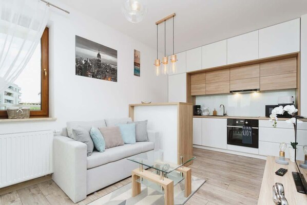 A bright, modern living room with a light-colored sofa, small coffee table, and open-plan design connected to a kitchen. Soft colors and minimalistic decor add a cozy touch.