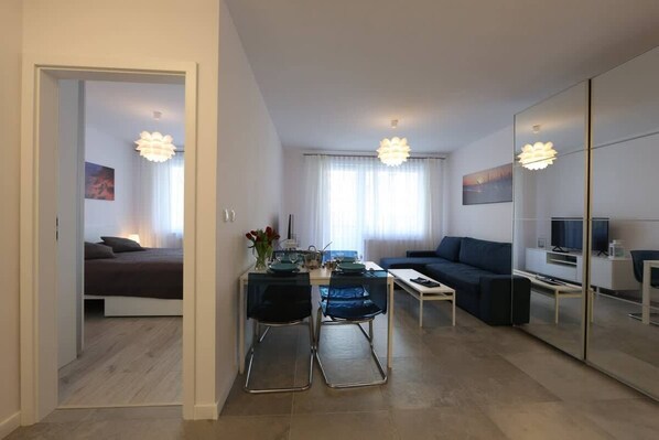 A modern living room featuring a sofa, dining table, and a chandelier, with bright natural light coming through large windows.