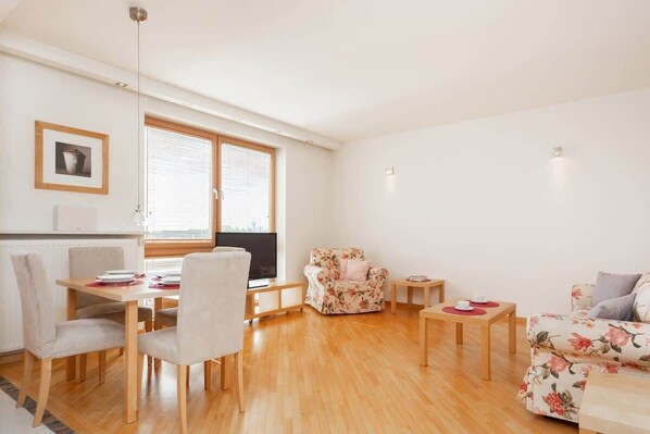 A spacious living room with a dining area, featuring wooden flooring, a cozy sofa bed, and a dining table for four. The large windows let in plenty of natural light.