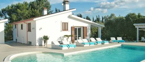 Pool and side view of villa with patio and sun loungers.