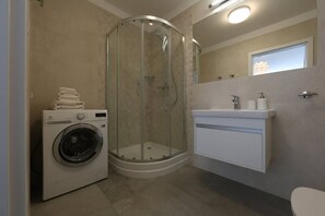 A compact bathroom featuring a glass-enclosed shower, a washing machine, and a white vanity, efficiently utilizing space.